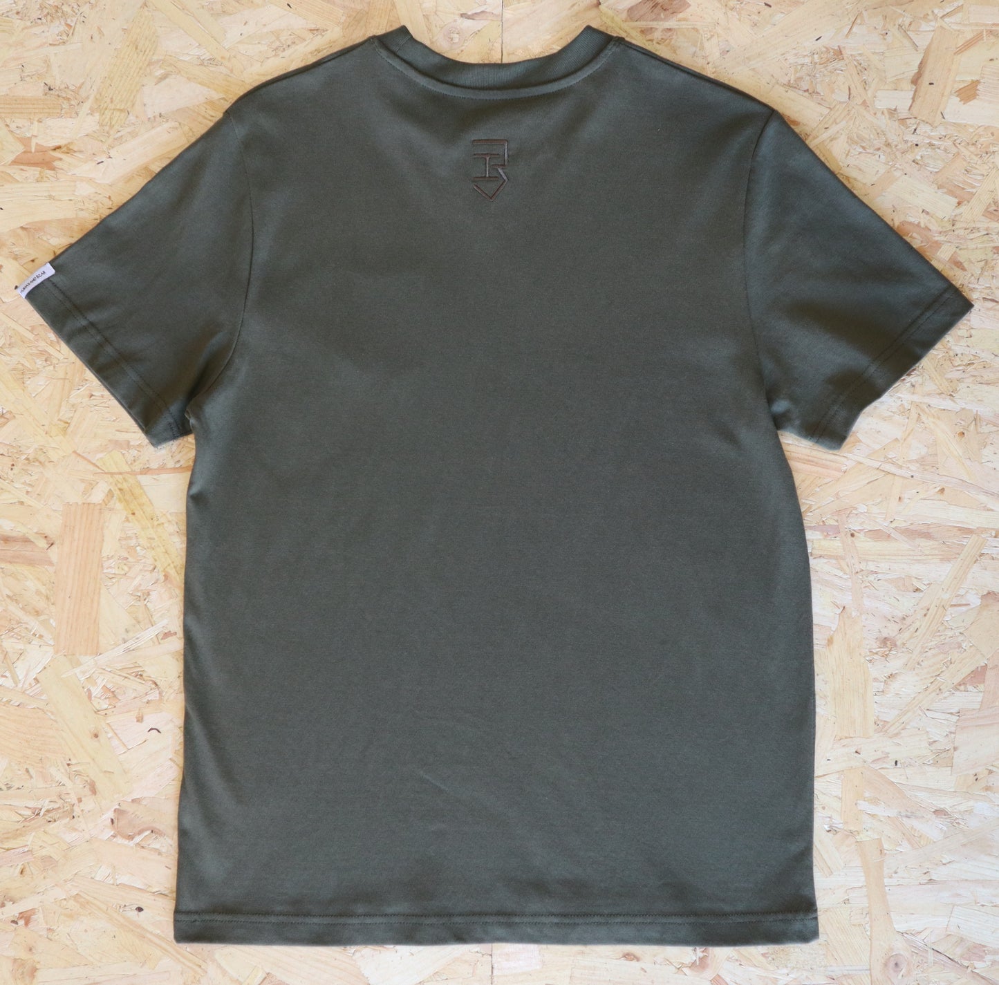 Khaki t-shirt with a logo on the back hem, placed on a wooden surface